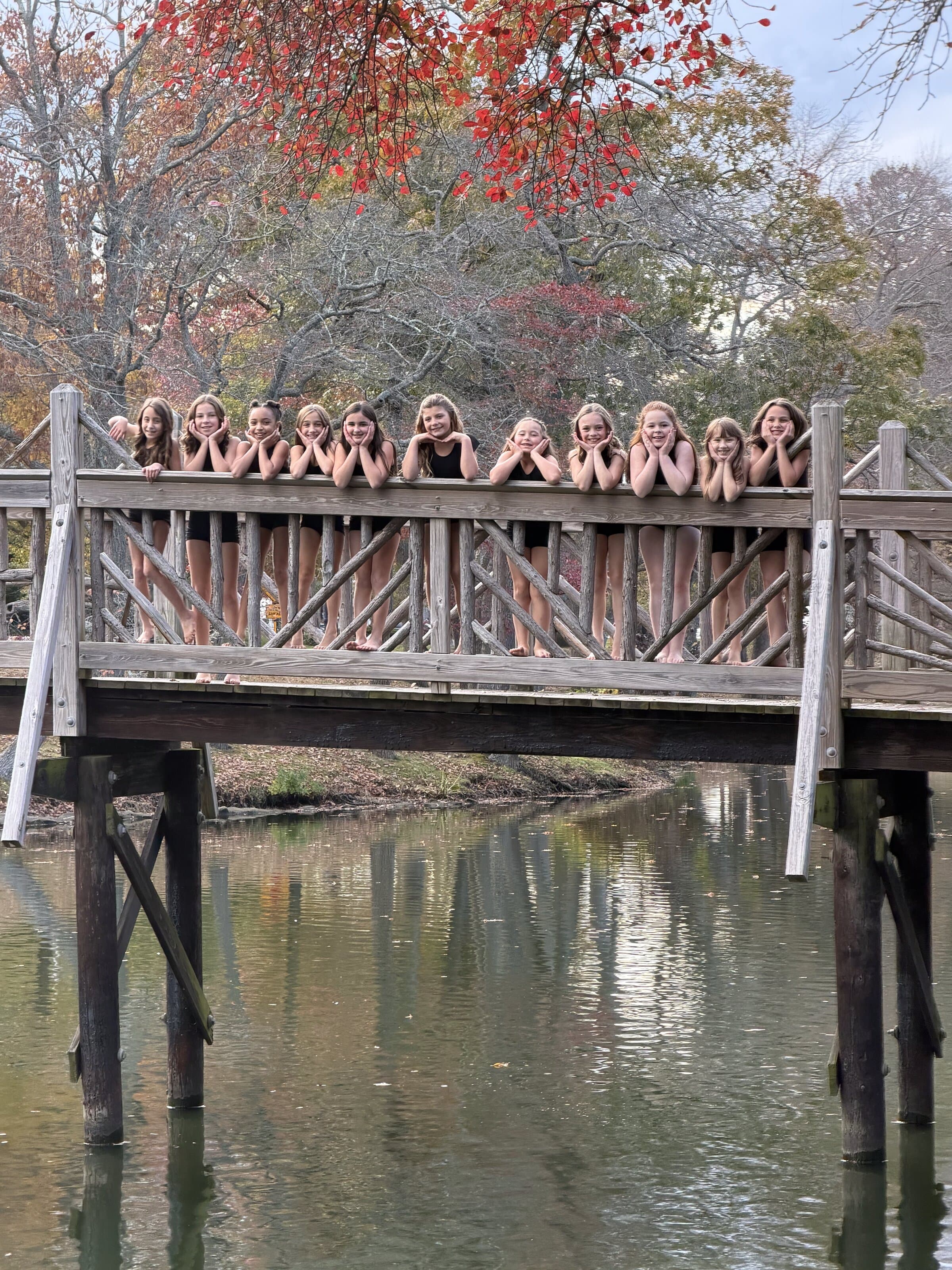 Dancers lined up smiling on a wooden bridge surrounded by fall foliage